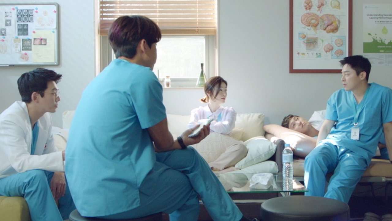 Medical professionals in scrubs engaged in a casual conversation in a hospital room. One doctor in a white coat is sitting on the left while three others, all in blue scrubs, are seated around a patient resting on a sofa. The room features medical charts on the wall and a calm, collaborative atmosphere.