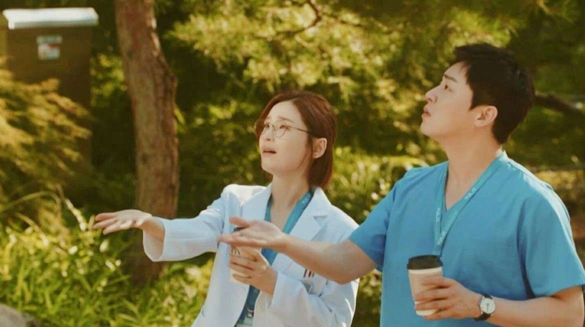 Two characters from 'Hospital Playlist+' enjoying a conversation outside, holding coffee cups and looking up towards the sky.