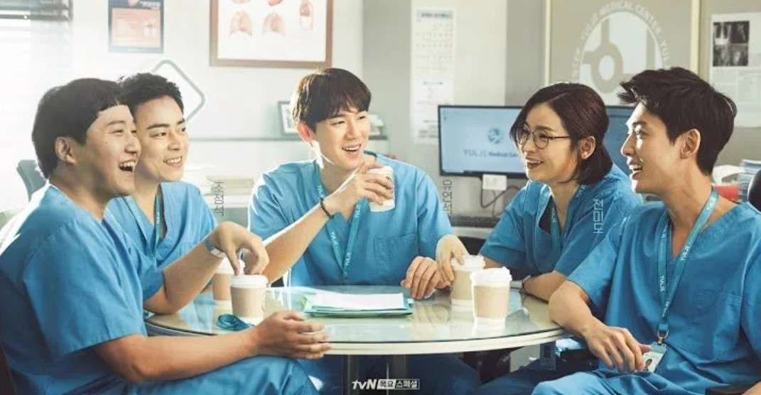 Cast of 'Hospital Playlist+' laughing and enjoying coffee together in a hospital setting, dressed in blue scrubs and sharing a lighthearted moment.