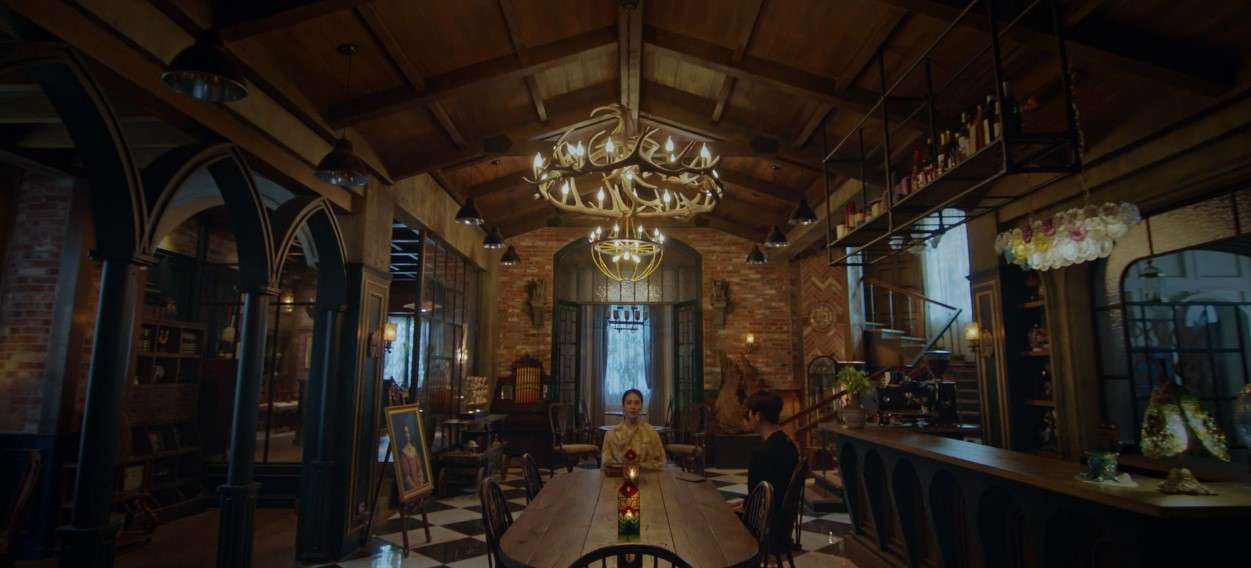 Interior scene from 'The Witch’s Diner' featuring two characters seated at a wooden table in a beautifully designed, mystical restaurant with vintage decor, high ceilings, and an elaborate chandelier, creating an atmospheric and enchanting setting.