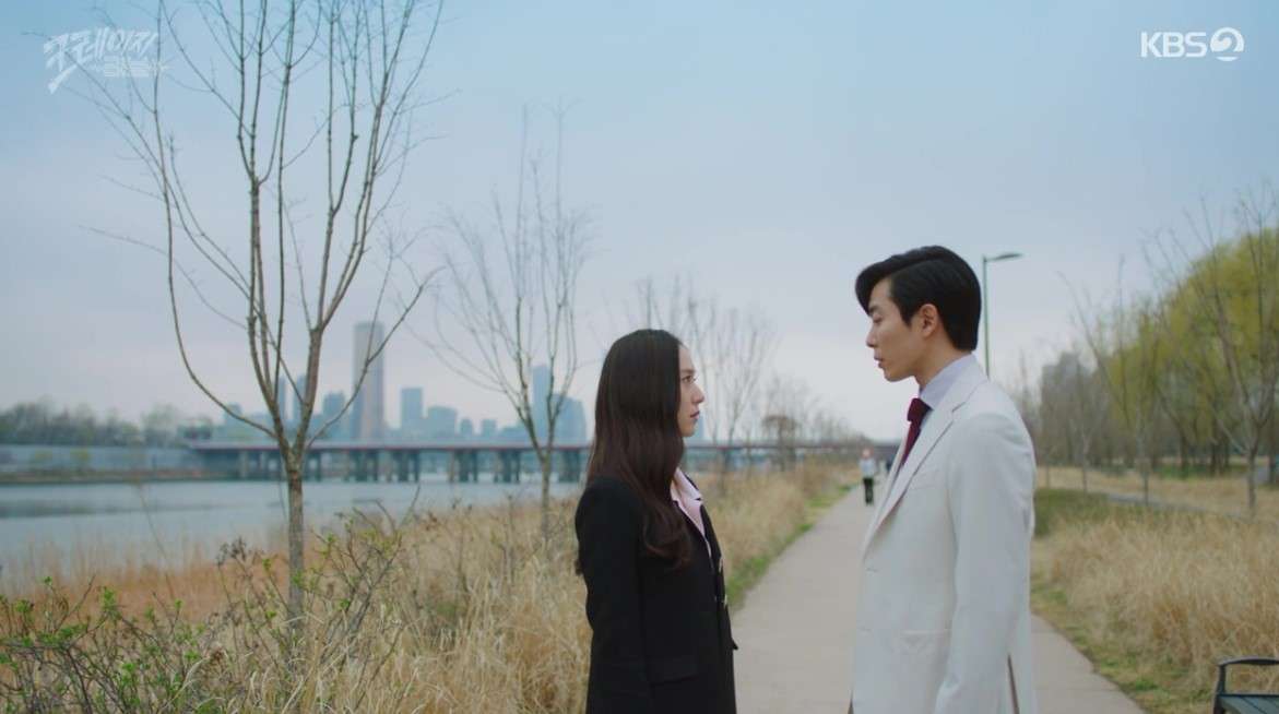 Scene from 'Crazy Love' showing the lead characters standing face-to-face in a scenic outdoor setting, with a city skyline in the background and trees along a path, capturing a tense and emotional moment.