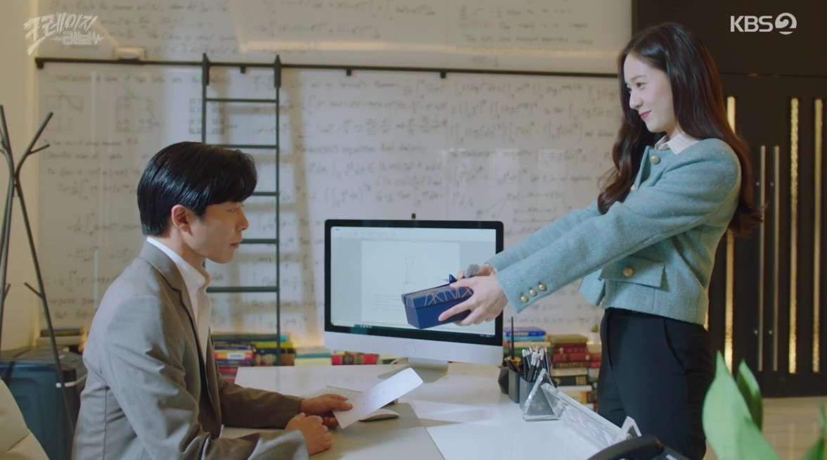 Scene from 'Crazy Love' showing the female lead offering a gift box to the male lead, who is seated at his desk in an office, with mathematical equations on the wall and a computer in the background.