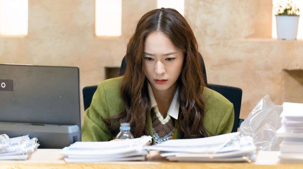 Scene from 'Crazy Love' showing the female lead working intensely at her desk, surrounded by paperwork, with a focused expression while sitting in an office setting.