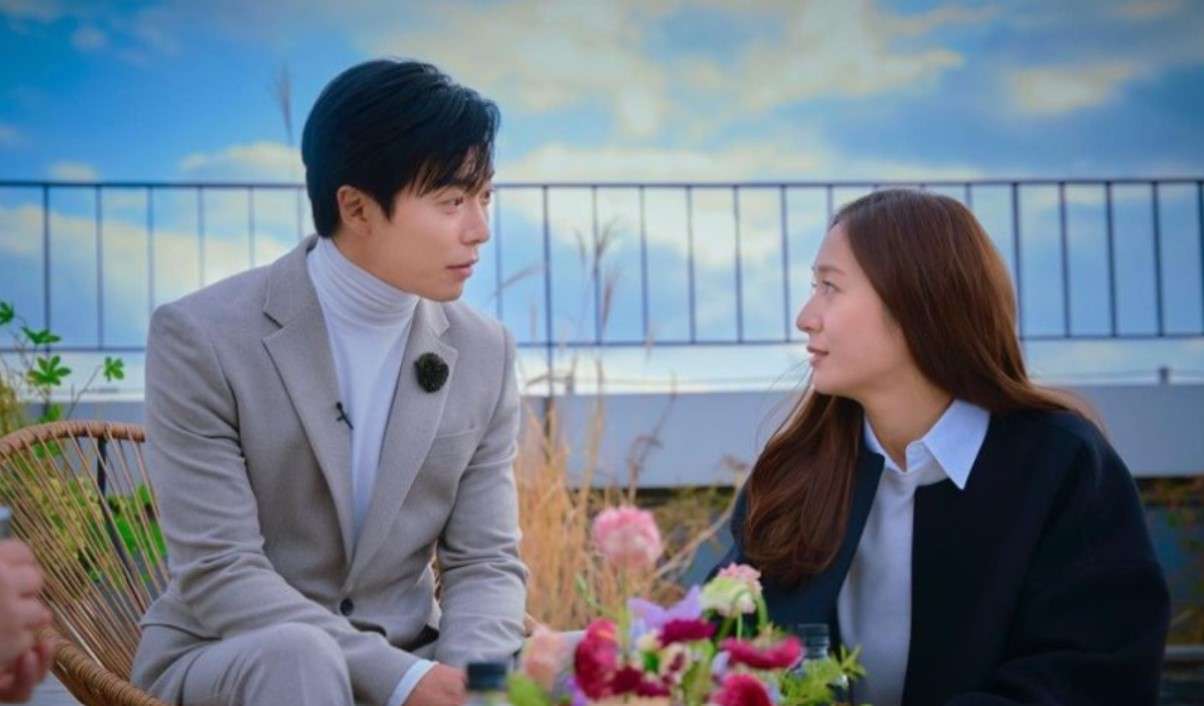 Scene from 'Crazy Love' featuring the male and female leads sitting across from each other, engaging in a deep conversation on a rooftop with a beautiful sky backdrop, surrounded by flowers.