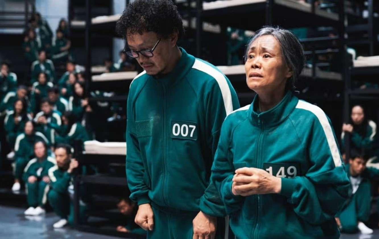  In a room, two characters in green uniforms are shown, representing a scene from Squid Game Season 3.