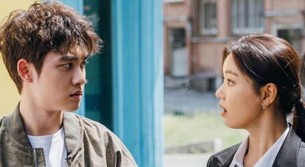 Close-up scene of two characters in casual and formal attire, engaged in a serious conversation, set against a colorful urban background with a building and greenery in the distance.