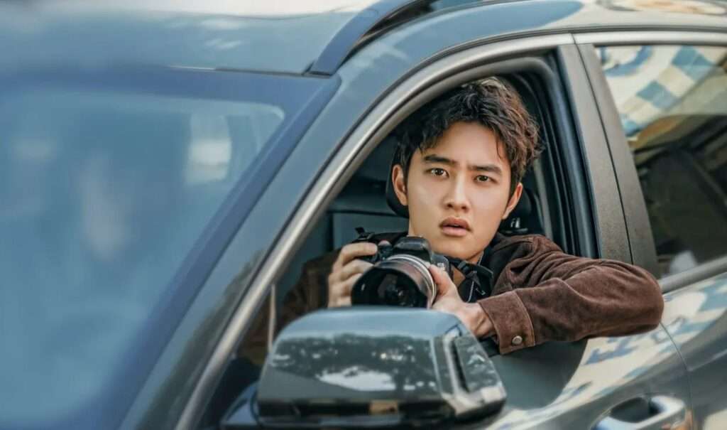 A character in a brown jacket holding a camera, leaning out of a car window, capturing a moment with a focused expression, set against a reflective vehicle surface and outdoor backdrop.