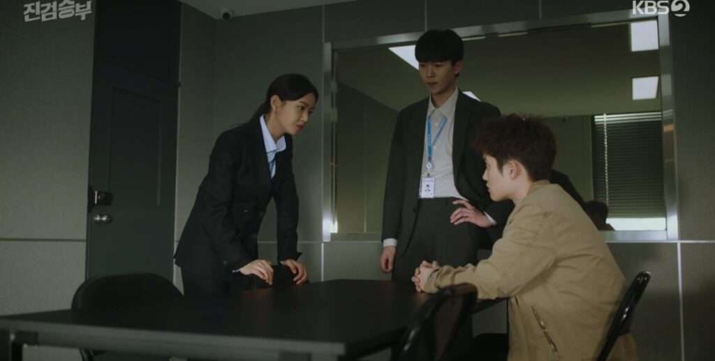Two men and a woman stand around a table, engaged in discussion, from the drama "Bad Prosecutor."