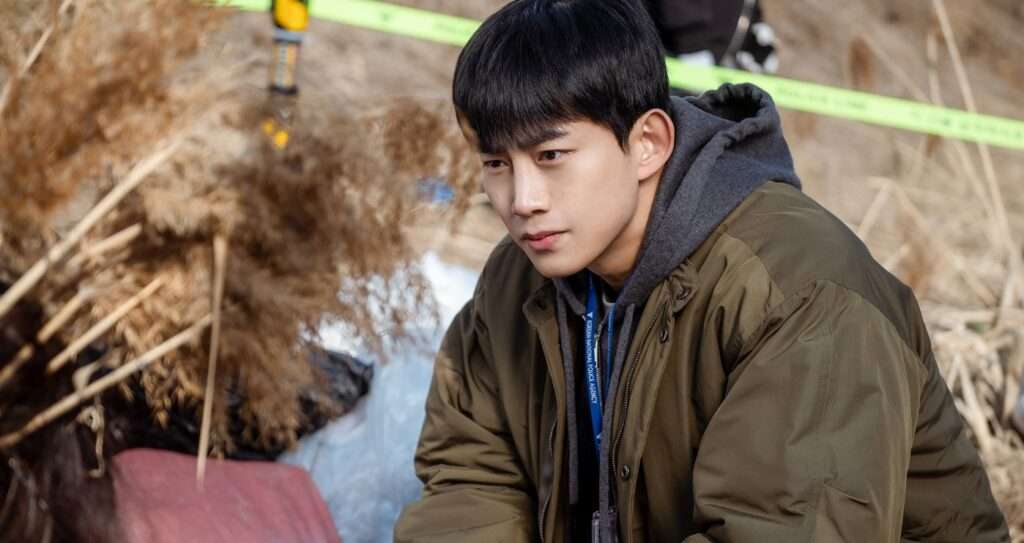 A scene from the Blind full review featuring a detective in a green jacket with a police badge, investigating a crime scene surrounded by yellow tape and dry grass, capturing the drama's intense atmosphere.