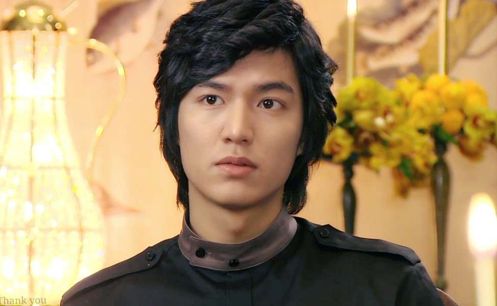 Lee Min-ho in a stylish black outfit, posing with a serious expression in an elegant setting with chandeliers and yellow flowers, a glimpse into his Lee Min-ho profile and biography from Boys Over Flowers.