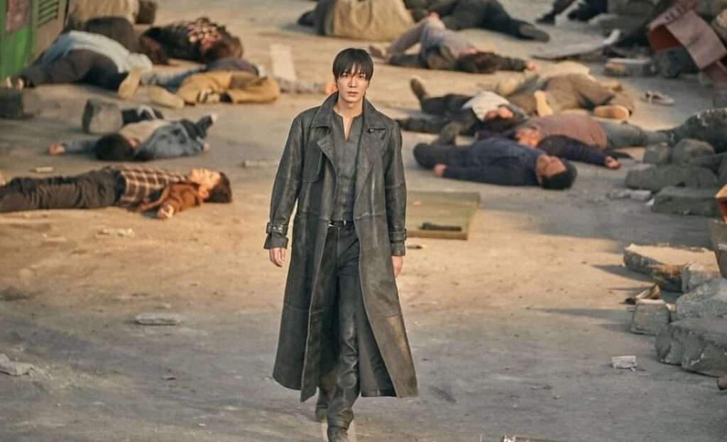 Lee Min Ho in a trench coat walks solemnly through a field scattered with lifeless bodies, evoking a haunting atmosphere. 