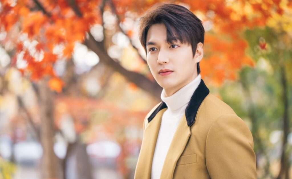 Lee Min Ho, wearing a yellow jacket and white shirt, poses in front of colorful autumn foliage.