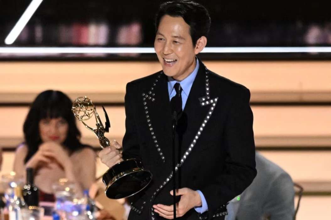 Lee Jung-jae smiling and holding an Emmy award on stage, dressed in a stylish black suit with silver embellishments.