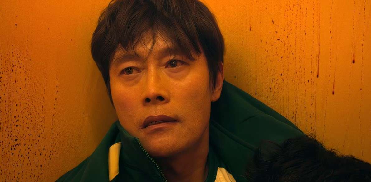 Lee Byung-hun in a green jacket, seated on a yellow wall, showcasing his relaxed demeanor in a vibrant setting.