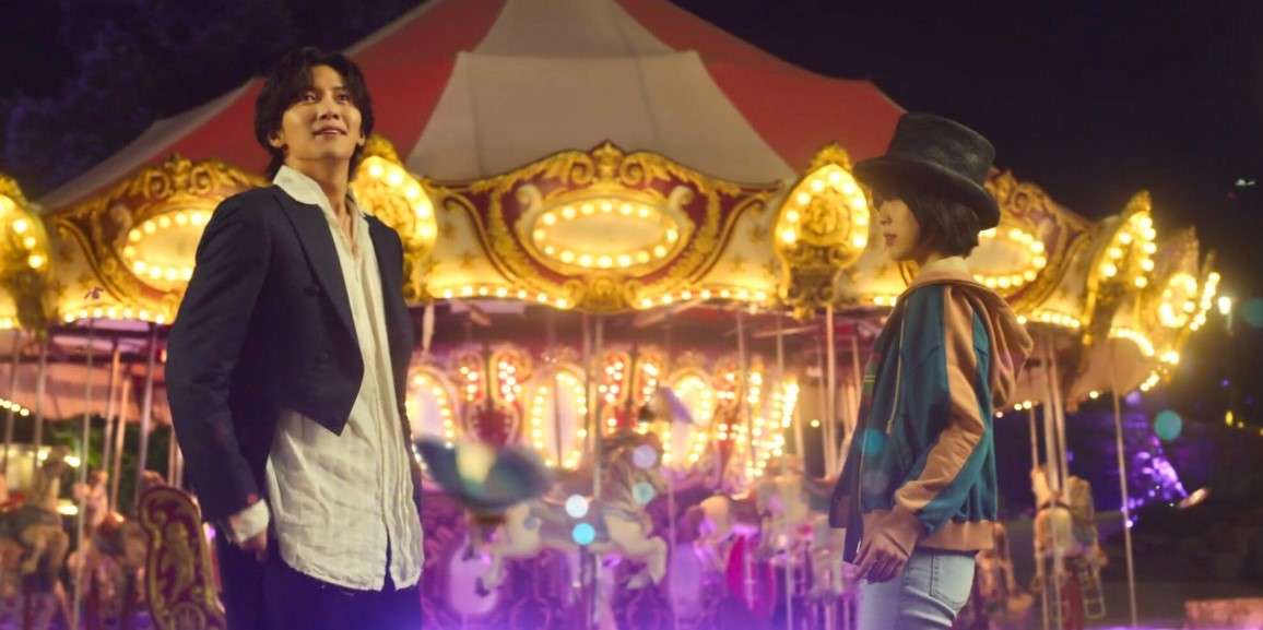Two characters from "The Sound of Magic" standing in front of a glowing carousel at night, one in a suit and the other in a hoodie and top hat.