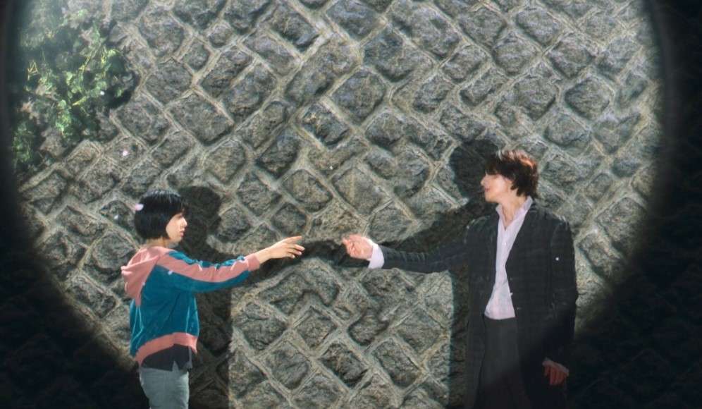 Scene from "The Sound of Magic" showing two characters reaching out to each other against a textured stone wall, casting dramatic shadows.
