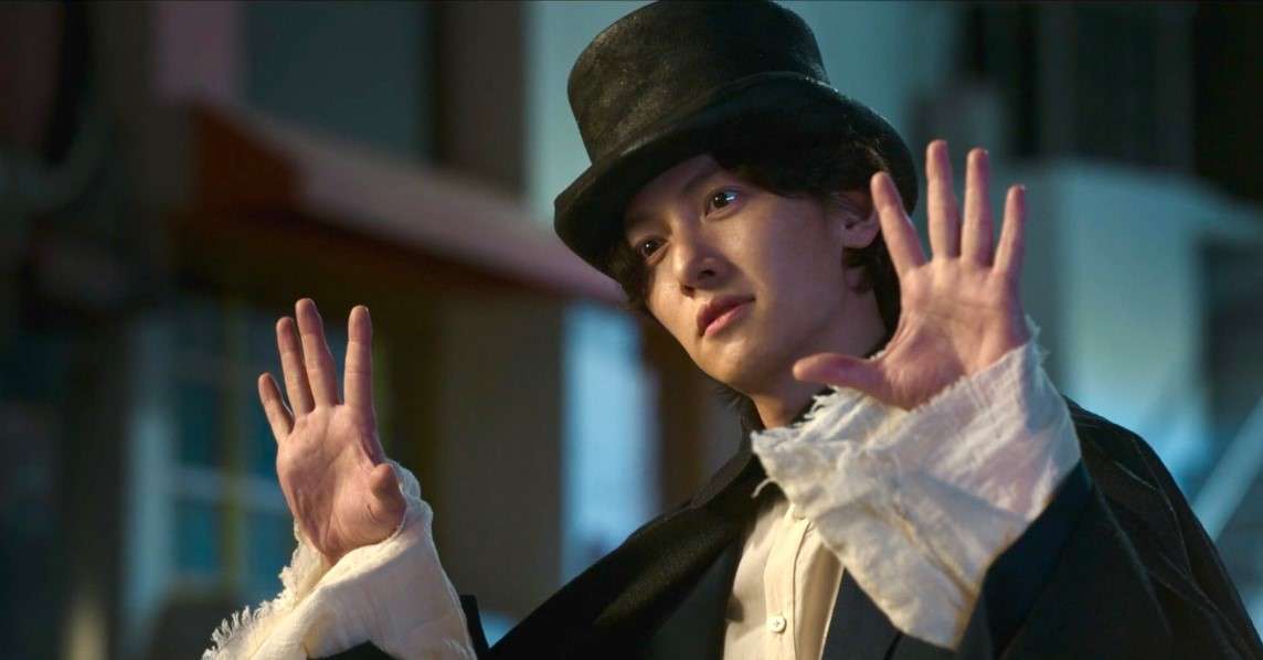 A character from "The Sound of Magic" in a top hat and black coat, raising hands in a magical gesture against a blurred urban background.