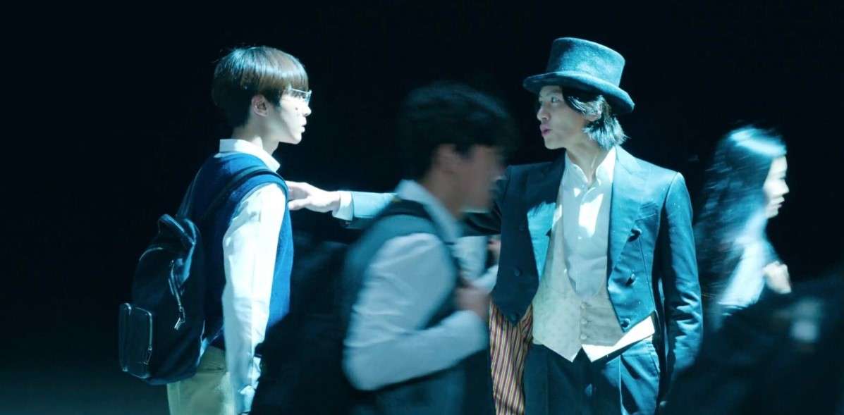 Scene from "The Sound of Magic" showing a student in a vest and backpack interacting with a character in a top hat and suit, set against a dark background.