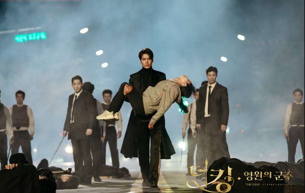 Dramatic scene from The King: Eternal Monarch with a man in a black coat carrying an injured person through a foggy street, surrounded by suited figures and fallen bodies, with Korean text and title overlay.