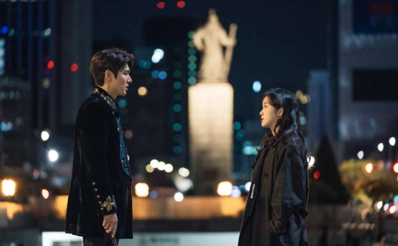 Romantic night scene of a couple facing each other in a cityscape, with a man in a black velvet jacket and a woman in a dark coat, standing near a statue with blurred city lights in the background.