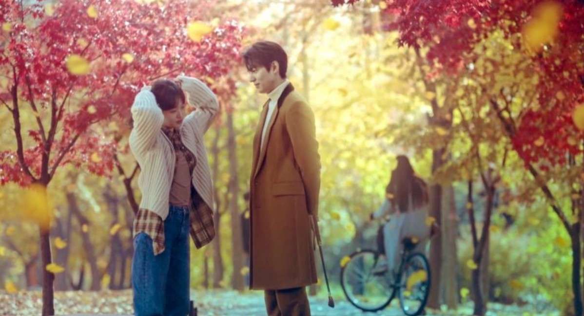 Romantic autumn scene with a couple standing under vibrant red and yellow trees, surrounded by falling leaves, while a person with a bicycle is visible in the background.