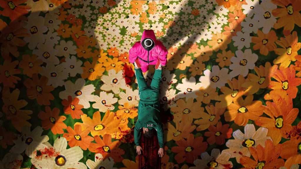 Overhead shot from Squid Game Season 3 showing a masked guard in a pink suit dragging a bloodied contestant in green uniform (Player 214) across a surreal, flower-patterned floor.